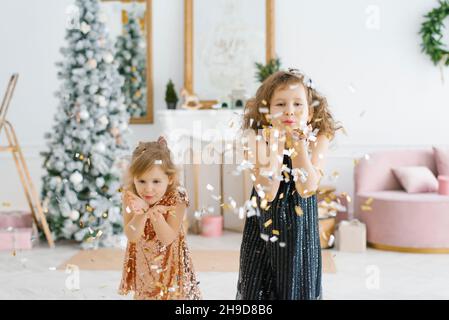 Zwei glückliche Kinder feiern zu Hause Neujahr und Heiligabend. Sie verwandeln sich in Konfetti, die in verschiedene Richtungen fliegen. Die Mädchen lächeln und Stockfoto