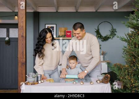 Mama, Papa und kleiner Sohn Rollen den Teig für Ingwerkekse oder Lebkuchenhaus aus. Vorbereitung auf Weihnachten Stockfoto