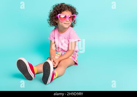 Portrait von attraktiven fröhlichen Mädchen tragen große rosa specs sitzen isoliert auf hellen blaugrün türkisfarbenen Hintergrund ruhen Stockfoto