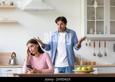 Unglückliche junge europäische Dame mit Smartphone ignoriert beleidigten wütenden schreienden Mann in der Küche Stockfoto