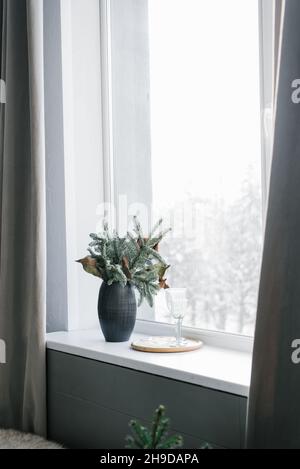 Weihnachtliche Komposition aus Christbaumzweigen in einer schwarzen Vase auf dem Fenster eines gemütlichen Hauses Stockfoto