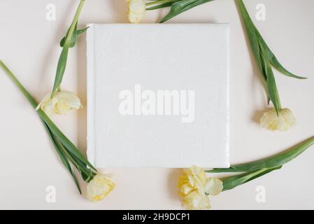 Weißes Familien- oder Hochzeitsfotoalbum mit Ledereinband mit leerem Platz für Text, umgeben von gelben Tulpen auf hellem Hintergrund. Familie Foto Archi Stockfoto