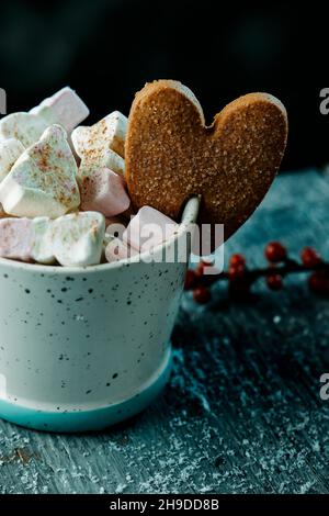 Nahaufnahme eines herzförmigen Lebkuchenkekse in einer Tasse Kaffee, gekrönt mit einigen verschiedenen Formen von Marschmalchen, neben einem Zweig von winterberry o Stockfoto