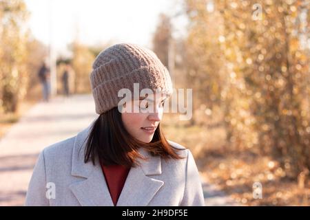 Lächelndes Mädchen mit Strickmütze und Mantel geht auf einem gepflasterten Weg in einen Herbstpark Stockfoto