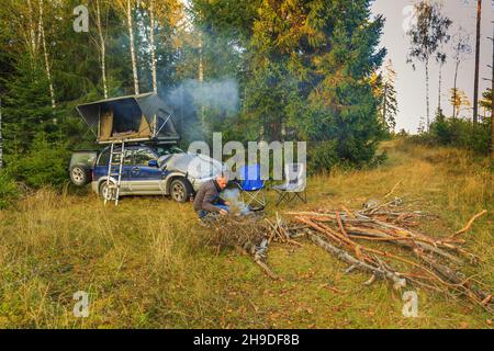 Der Mann zeltet mit einem wilden Auto mit dem Dachzelt Jimba Jimba von Sheepie im Nadelwald und bereitet auf dem rauchenden Holzfeuer in einer Bratpfanne Essen zu Stockfoto