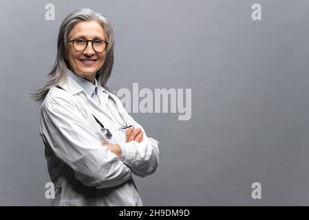 Kopfbild einer intelligenten professionellen reifen Ärztin mit grauem Haar, Brille, medizinischem weißem Kleid und Stethoskop, Blick auf die Kamera, Therapeut Arzt isoliert auf grau stehend Stockfoto