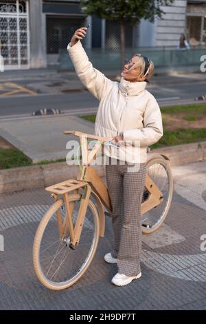 Ein großer Winkel der erwachsenen Frau mit einem Öko-Fahrrad aus Holz lächelt und nimmt Selfie auf, während sie auf dem Bürgersteig auf der Stadtstraße steht. Stockfoto