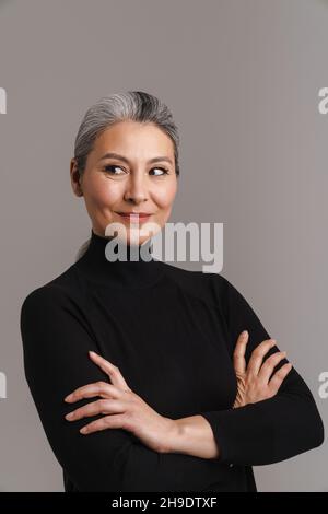 Reife weißhaarige Frau lächelt, während sie sich mit verschränkten Armen auf grauem Hintergrund posiert Stockfoto