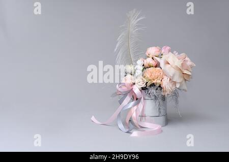 Elegante Blumenkomposition mit Feder, kleinen Cupid- und Pastellbändern in zylindrischer Geschenkbox. Zartes Bouquet von Elfenbeinrosen auf grauem Hintergrund. Stockfoto