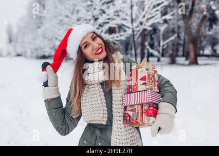 Glückliche Frau hält Weihnachtsgeschenke Geschenkboxen im verschneiten Winterpark tragen Weihnachtsmütze im Freien. Festliche Feiertage. Mädchen trägt warm gestrickt c Stockfoto