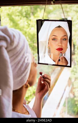 Schöne junge Frau, die roten Lippenstift aufgesetzt hat, während sie am Fenster steht und auf den Spiegel schaut. Stockfoto