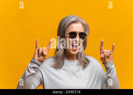 Verrückte positive sanguine ältere Frau in Sonnenbrillen zeigt Zeichen von Hörnern, lustig urkomisch reifen modernen grauhaarigen Dame machen Symbole des Felsens stehend isoliert über gelbem Hintergrund Stockfoto