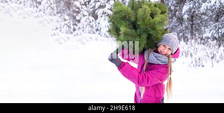 Ein fröhliches Mädchen, 10-11 Jahre alt, mit Strickmütze und Schal, das einen Weihnachtsbaum hält, lächelt und blickt auf die Kamera gegen den Schnee und die Bäume. Winter Stockfoto