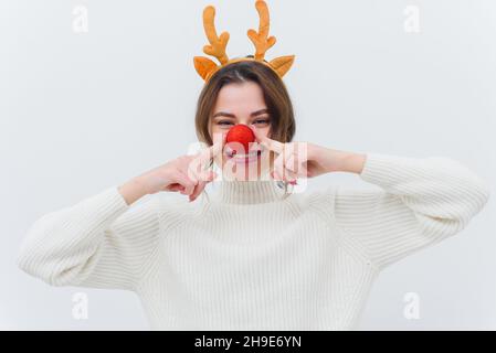 Foto einer überfröhlichen lustigen Dame in einem weihnachtlichen Hirsch-Kostüm und einem weißen Pullover mit roter Kugel und weißem Hintergrund. Stockfoto