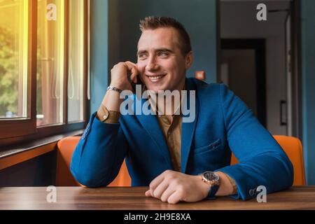 Ein stilvolles, lächelndes, positives Portrait eines europäischen Geschäftsmannes sitzt in einer blauen Jacke an einem Tisch in einem Café und spricht auf einem Smartphone lo Stockfoto