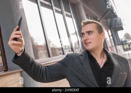 Geschäftsmann Mann von schönen europäischen Auftritt, in einer Jacke und Hemd macht ein Selfie oder spricht auf einem Videoanruf auf einem Smartphone auf der Straße draußen. Stockfoto