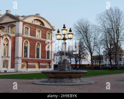 London, Greater London, England, 04 2021. Dezember: Lampen leuchten in der Abenddämmerung auf einem Brunnen in Greenwich zur Weihnachtszeit mit einem festlichen Licht dahinter. Stockfoto