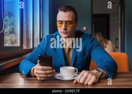 Ein seriöser Geschäftsmann mit europäischem Auftritt in Sonnenbrillen, eine blaue Jacke sitzt in einer Kaffeepause an einem Tisch in einem Café, hält ein Smartphone oder Handy Stockfoto