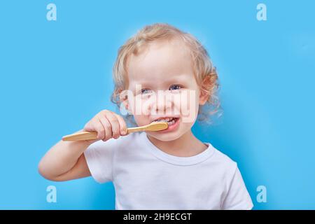 Das lockige Mädchen putzt ihre Zähne mit einer hölzernen Zahnbürste auf einem blauen Hintergrund mit Platz für Kopien. Das Konzept des Recyclings und umweltfreundlich. Stockfoto