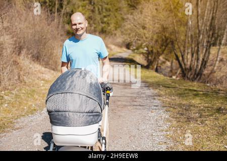 Junger Mann in Cyan T-Shirt schieben Baby-Wagen auf Waldstraße im Frühjahr Stockfoto