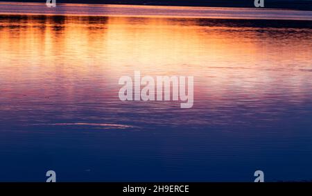 Abstraktes Küstenmotiv mit Langzeitbelichtung für intensiven Farbhintergrund mit orangefarbenem Farbton, der einen Hauch von Landsilhouette und Wolken am Horizont zeigt. Stockfoto