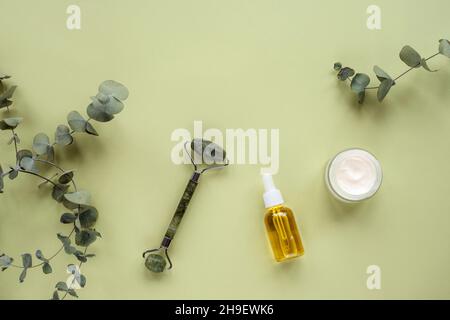 Kosmetisches Serum in Glasflasche, Cremedose, Gesichtswalze und Eukalyptusblättern auf hellgrünem Hintergrund. Hautpflege, natürliche Bio-Kosmetik-Konzept. T Stockfoto