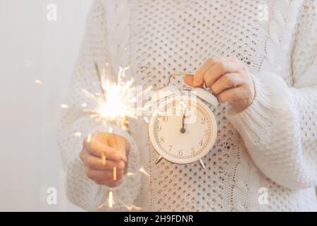 Ein Mädchen in weißem Pullover hält Wunderkerzen, Uhr ist ein Wecker Symbol des neuen Jahres, Pfeile um Mitternacht 12 Uhr. Stockfoto