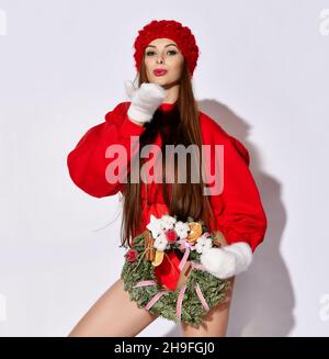 Fröhliches Mädchen in roten kurzen Oberteil Pullover, Body, Hut und Fäustlinge mit dekorierten Weihnachtskranz in der Hand sendet einen Kuss Stockfoto