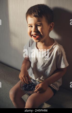 Kaukasischer Junge, der aus einer Schüssel frische Blaubeeren isst und lächelt und auf dem Sofa sitzt, während er die Kamera anschaut. Stockfoto