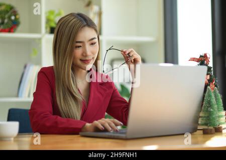 Elegante asiatische junge Geschäftsfrau in rotem Anzug, die eine Brille hält, während sie am Schreibtisch am Laptop arbeitet. Stockfoto