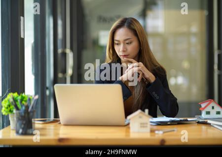 Asiatische junge Immobilienmaklerin oder Geschäftsfrau konzentrierte sich auf ihr Projekt auf Laptop-Computer. Stockfoto