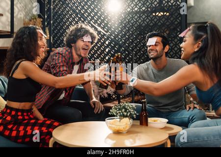 Eine Gruppe glücklicher männlicher und weiblicher Freunde, die Namensspiel spielen und mit einer Bierflasche anstoßen, während sie zu Hause während der Hausarming-Party sitzen Stockfoto