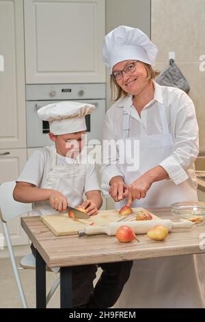 Mutter und Sohn in weißen Chefkleidern schnitten in ihrer eigenen Küche mit einem Messer Birnen. Eine Erwachsene Frau mit einem kleinen Jungen wird einen Kuchen kochen Stockfoto