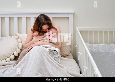 Eine Mutter küsst und tröstet einen weinenden kleinen Jungen, der auf dem Bett sitzt Stockfoto