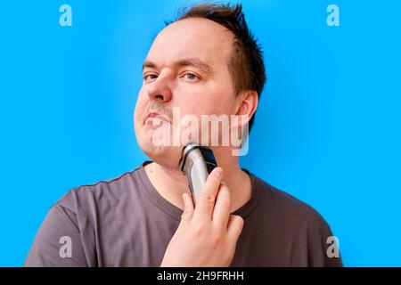Porträt eines zerzausten erwachsenen Mannes, der mit einem elektrischen Rasiermesser Stoppeln rasiert, blauer Studiohintergrund Stockfoto