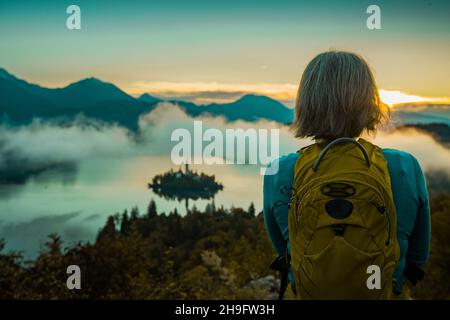 Kurzhaarige Wandererin wartet und beobachtet den Morgensonnenaufgang über dem See von Bled und der Insel darauf. Stockfoto