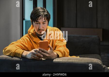 Ein Asiat sitzt in einem kleinen Büro auf dem Sofa und kommuniziert per Mobiltelefon. Das Konzept von Kleinunternehmen und Online-Kommunikation. Stockfoto