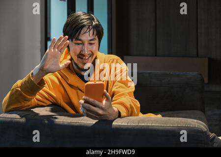 Ein Asiat sitzt in einem kleinen Büro auf dem Sofa und kommuniziert per Mobiltelefon. Das Konzept von Kleinunternehmen und Online-Kommunikation. Stockfoto