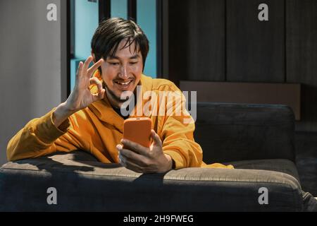 Ein Asiat sitzt in einem kleinen Büro auf dem Sofa und kommuniziert per Mobiltelefon. Das Konzept von Kleinunternehmen und Online-Kommunikation. Stockfoto