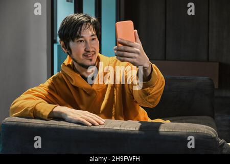 Ein Asiat sitzt in einem kleinen Büro auf dem Sofa und kommuniziert per Mobiltelefon. Das Konzept von Kleinunternehmen und Online-Kommunikation. Stockfoto