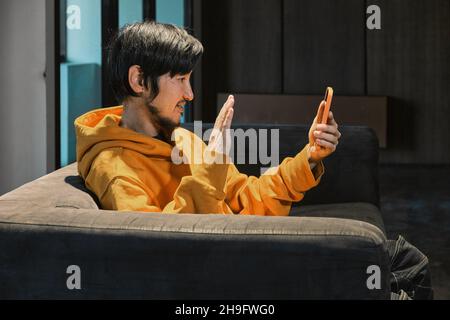 Ein Asiat sitzt in einem kleinen Büro auf dem Sofa und kommuniziert per Mobiltelefon. Das Konzept von Kleinunternehmen und Online-Kommunikation. Stockfoto