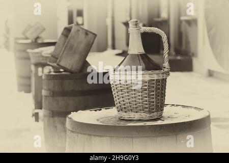 Flasche Rotwein im Korbkorb. Große Vintage-Weinflaschen mit Rotwein, die auf einem Fass stehen. Stockfoto