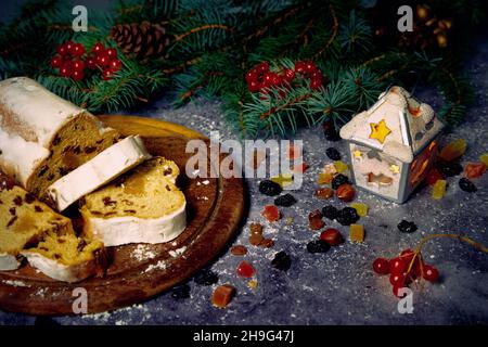 Chirstmass-Komposition. Stollen auf einem Holztablett, kandierte Früchte, Rosinen und Viburnum-Beeren mit Christbaumzweigen auf geklemmtem Hintergrund. Stockfoto