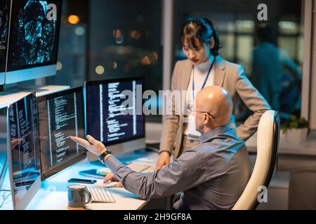 Zwei interkulturelle Kollegen interagieren vor Computermonitoren mit kodierten Daten, während einer von ihnen eine Präsentation macht Stockfoto