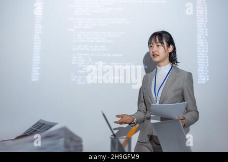 Junge erfolgreiche asiatische Trainerin mit Papier, die während der Präsentation auf dem Seminar mit einer Kollegin interagiert Stockfoto