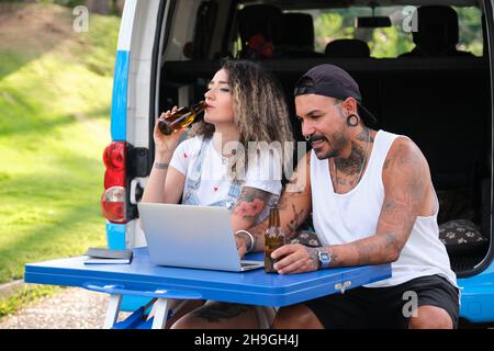 Das junge tätowierte Paar lächelt, trinkt Bier und benutzt den Laptop. Stockfoto