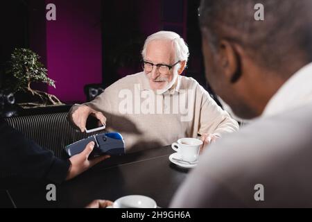 Älterer Mann in einer Brille, der mit dem Mobiltelefon über das Terminal bezahlte, in der Nähe eines verschwommenen afroamerikanischen Freundes Stockfoto