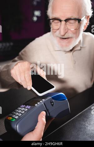 Älterer Mann in einer Brille, der sein Smartphone mit leerem Bildschirm benutzte, während er über das Terminal zahlte Stockfoto
