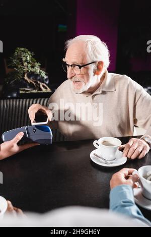 Älterer Mann in der Brille, der mit dem Handy über das Terminal in der Bar bezahlt hat Stockfoto