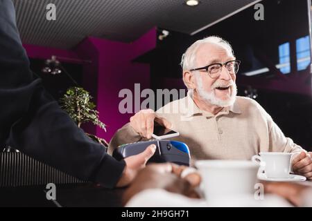 Älterer Mann in Brillen, der mit dem Smartphone über das Zahlungsterminal in der Kneipe bezahlt Stockfoto